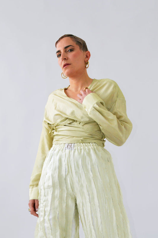 Femme portant la BLOUSE FLIP FLAP en soie verte citron, manches longues et design cache-cœur croisé, associée à un pantalon fluide clair, look élégant et lumineux.