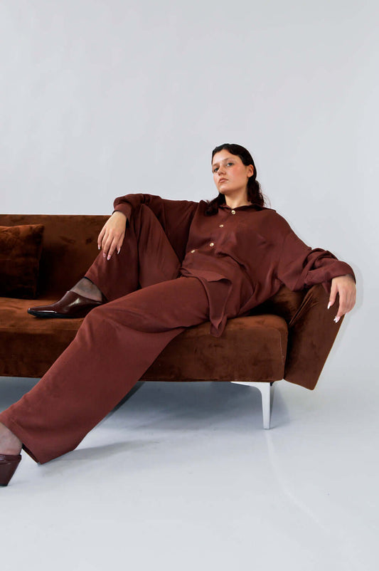 Modèle portant le PANTALON TOC TOC en viscose aubergine marron, assise sur un canapé marron, look chic et décontracté.