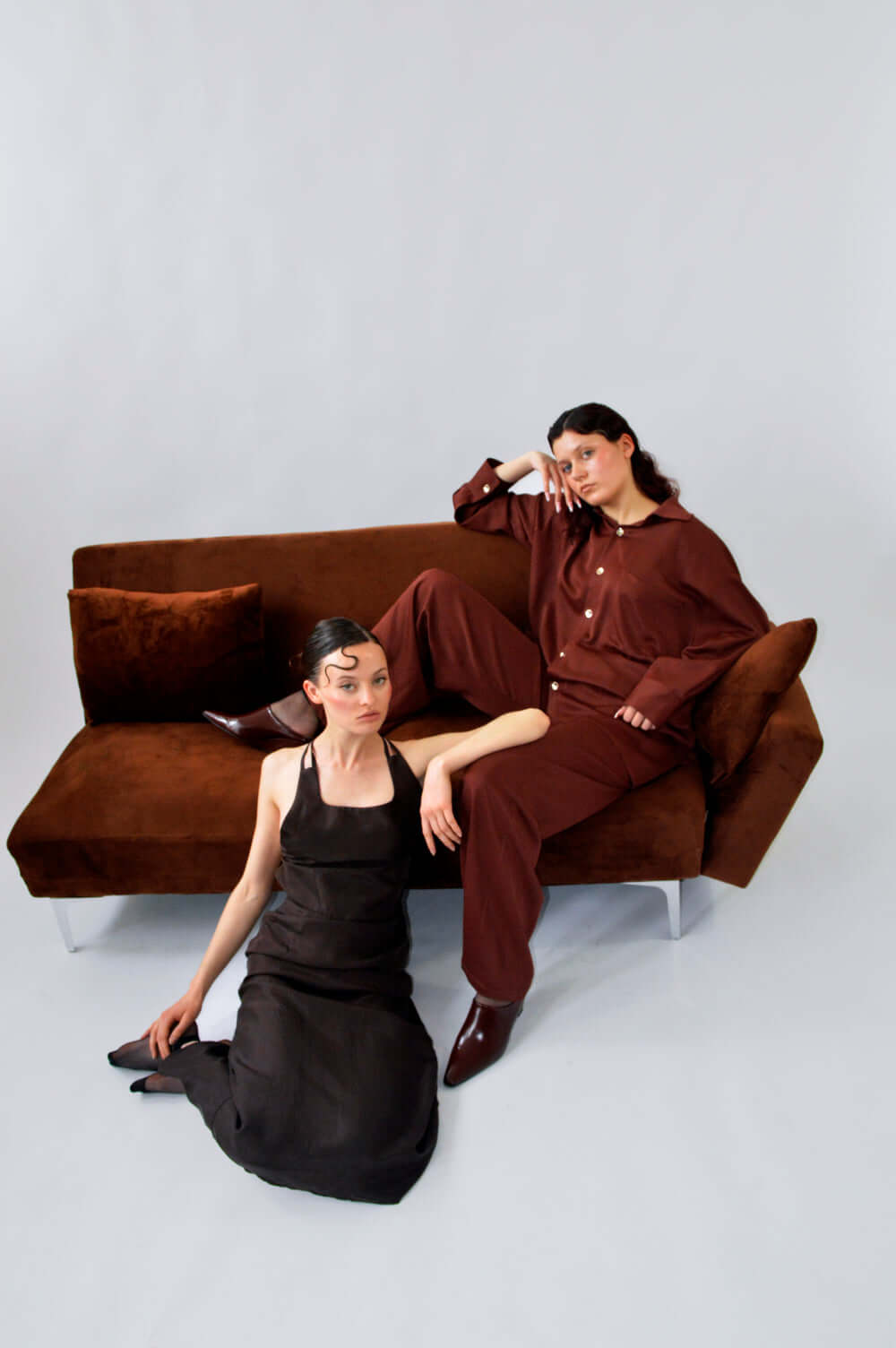 Deux modèles posant sur un canapé, l’un portant le PANTALON TOC TOC en viscose aubergine marron et l’autre la robe DING DONG.