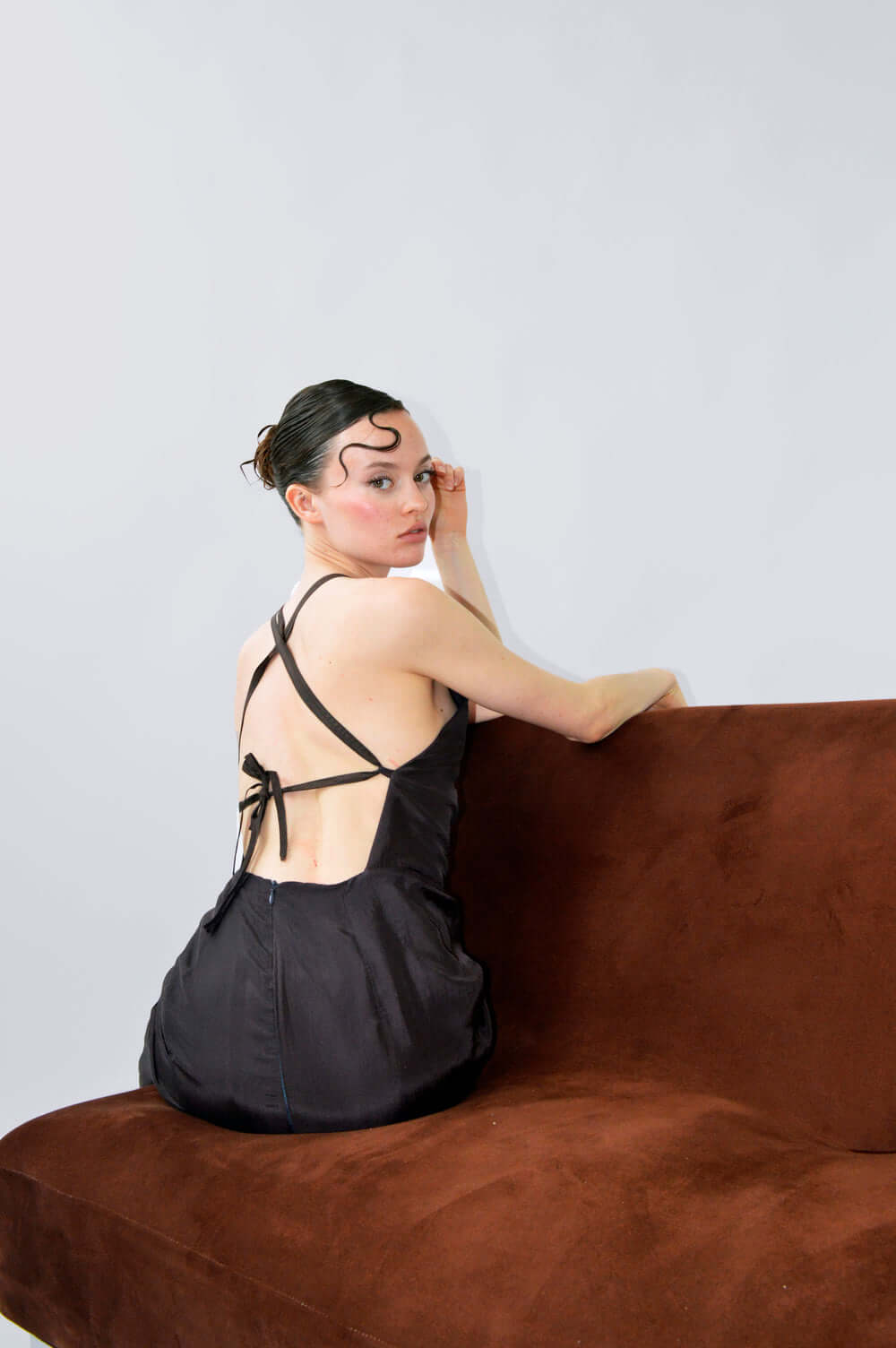 Modèle portant la robe DING DONG, dos nu avec son design minimaliste, assise sur un canapé marron.