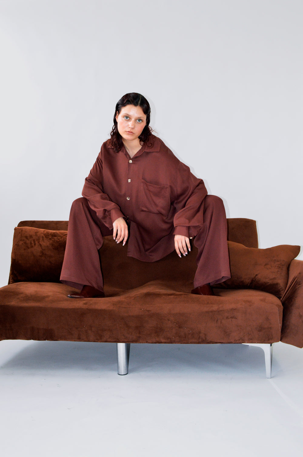 Modèle sur un canapé en velours brun, portant la CHEMISE TOC TOC en viscose lie de vin avec un pantalon assorti, illustrant une silhouette ample et audacieuse. 