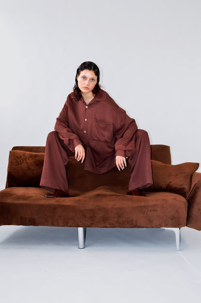 Modèle sur un canapé en velours brun, portant la CHEMISE TOC TOC en viscose lie de vin avec un pantalon assorti, illustrant une silhouette ample et audacieuse. 