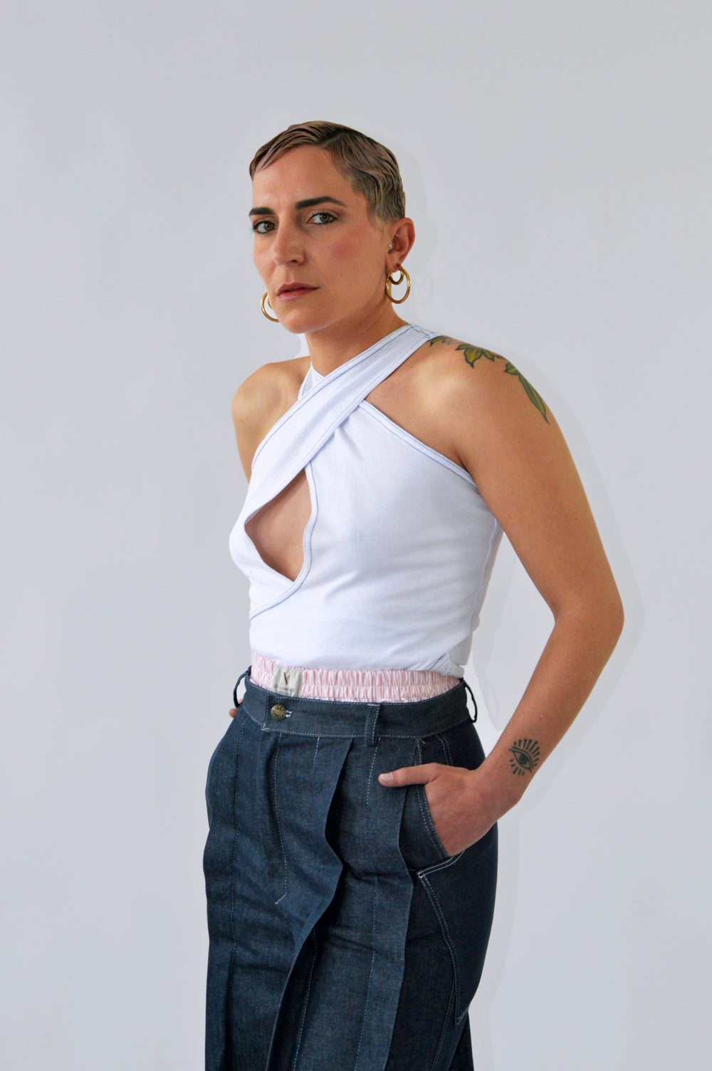 Femme portant le débardeur blanc croisé avec surpiqûres bleues de l’ensemble CLIC CLAC TOP, les mains dans les poches de son jean, pour un look décontracté et moderne.